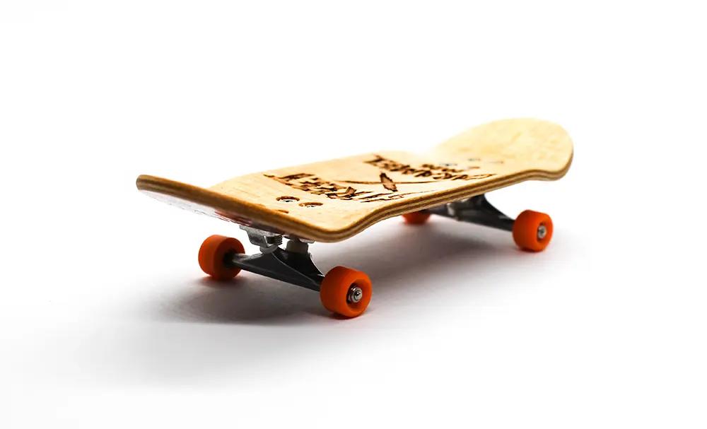 Light wood Street x Caramel Warrior Woman 34mm fingerboard with orange wheels and black trucks on a white background.